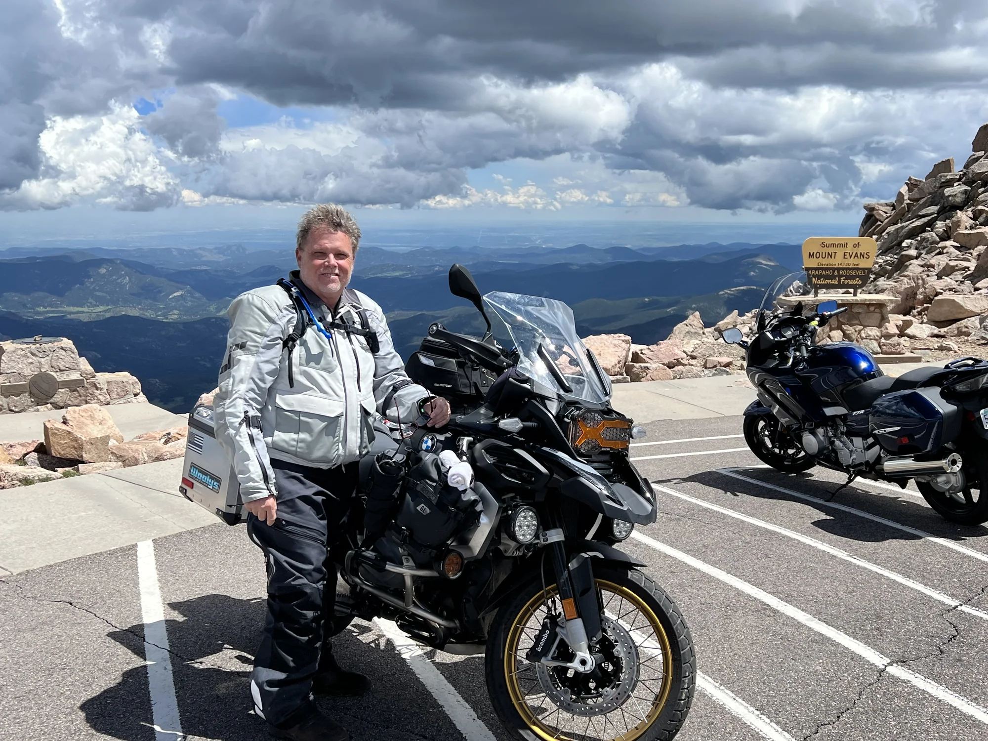 Paul Larson beside his BMW adventure motorcycle with the Summit of Mount Evans sign visible in Colorado.